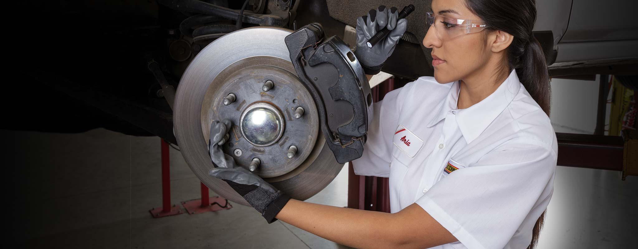 A Les Schwab technician inspecting a vehicle's brakes with a flashlight and protective glasses