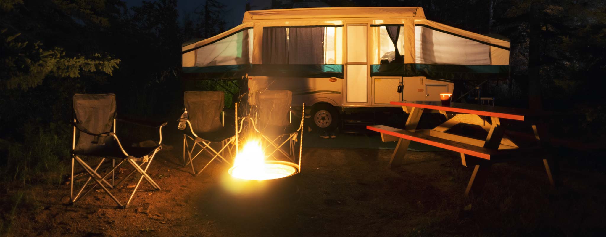 A pop-up tent trailer with it's lights on next to a campfire.